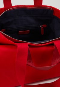Bolso rojo con material exterior liso y asas de tela. Cuenta con un interior azul marino, un bolsillo con cremallera y un cierre de cremallera plateado.
