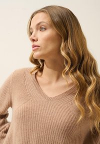 Sweter z dekoltem w serek, z długimi rękawami, w jasnobeżowym kolorze, wykonany z dzianiny w prążki, z teksturowanymi akcentami i luźnym krojem, noszony przez osobę z falującymi włosami.
