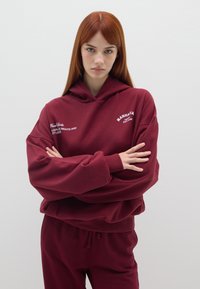 Felpa color bordeaux con una vestibilità comoda, dotata di cappuccio regolabile con lacci, tasca frontale e testo ricamato. Abbinata a pantaloni coordinati.