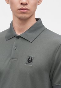 Άνδρας που φοράει γκρι πόλο μπλούζα Belstaff με γιακά και δύο κουμπιά, που δείχνει το λογότυπο στο αριστερό στήθος.