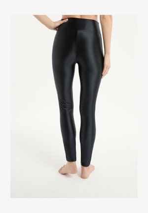 Mujer con leggins negros brillantes de cintura alta y puntos de agarre texturizados en pantorrillas y tobillos, de pie descalza contra un fondo liso.