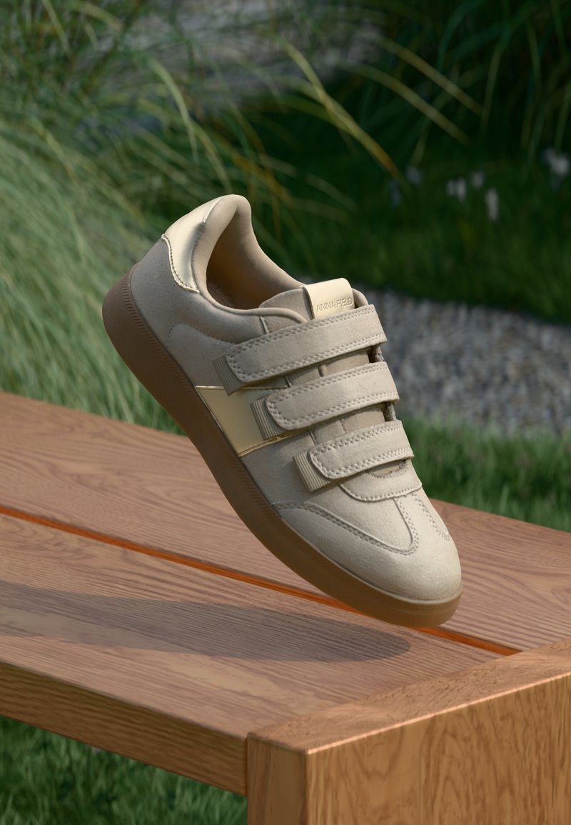 Baskets beiges avec trois bandes Velcro, en matériau suédé synthétique lisse, à bout rond et avec une semelle en caoutchouc, présentées sur une surface en bois.