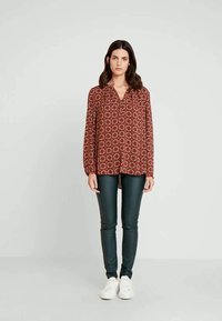 Blusa vermelha estampada com decote em V e mangas longas, combinada com calças justas verde-escuras e ténis brancos. Textura suave por toda a peça.