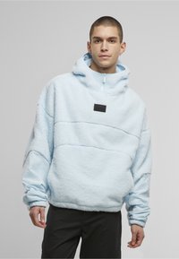 Junger Mann trägt einen hellblauen Fleecepullover mit Kapuze und elastischen Bündchen sowie schwarze Hose, steht vor einem schlichten grauen Hintergrund.