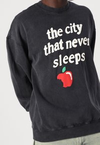 Sudadera de color gris oscuro con un llamativo texto en blanco que dice "la ciudad que nunca duerme" y un gráfico de una manzana roja con un mordisco.