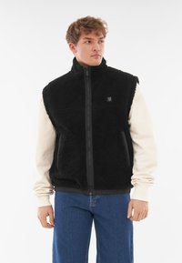 Fekete gyapjú mellény magas gallérral, cipzáras záródással és oldalsó zsebekkel. Karamell színű, hosszú ujjú ing fölött viselve és kék farmernadrággal párosítva.