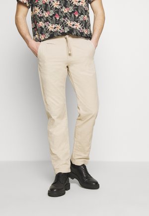 Hombre con pantalones beige con cordón, zapatos negros con cordones y una camisa negra de manga corta con flores, con las manos en los bolsillos.