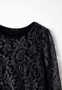 Svart långärmad topp med blommönstrad spetsöverdrag och enfärgad svart halsringning på vit bakgrund.