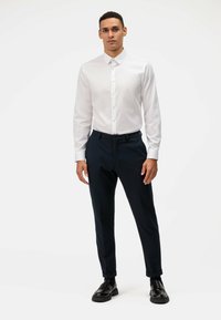 Witte blouse met lange mouwen en knoopsluiting, gecombineerd met donkere marineblauwe broeken. Model staat in zwarte schoenen tegen een effen achtergrond.