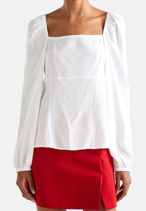 Haut blanc à manches longues avec des manches bouffantes, décolleté carré et tissu texturé. Porté avec une jupe rouge avec une fente sur le côté.