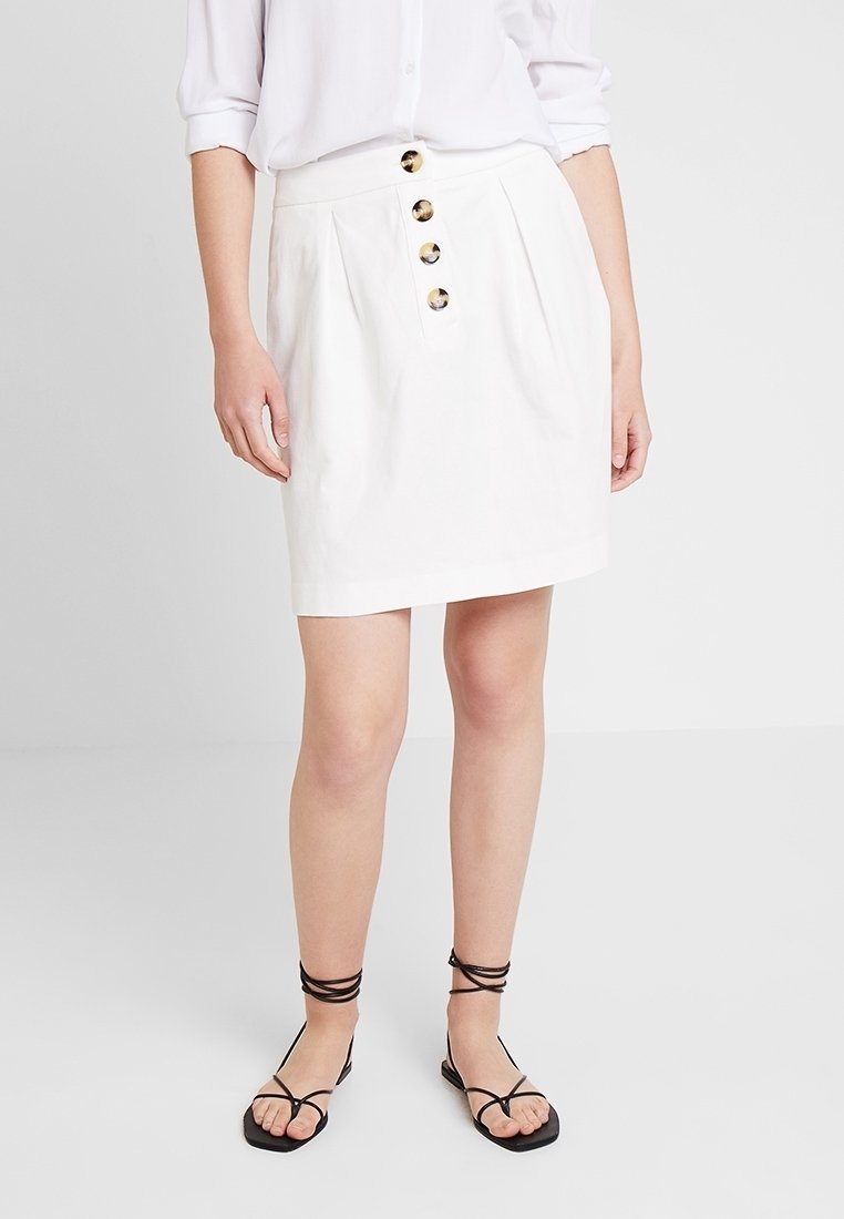 Valkoinen puuvillahame, jossa on laskokset ja viisi suurta kullanväristä nappia edessä. Yhdistetty mustiin remmikenkiin.