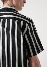Chemise à manches courtes noire avec des rayures verticales en vert clair et blanc. Elle présente un col classique et une coupe décontractée. Texture de tissu lisse.