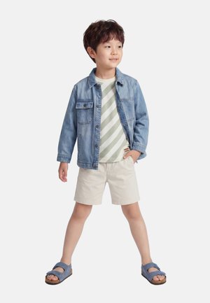 Jeune garçon debout, jambes écartées, vêtu d'une veste en jean bleue, d'un t-shirt rayé, de shorts beiges et de sandales bleues, regardant légèrement sur le côté.