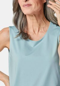 Top senza maniche di colore azzurro chiaro con una texture liscia e leggermente lucida e un collo rotondo. Il tessuto cade delicatamente sulle spalle.