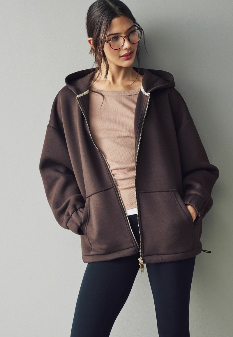 Brun lynlås hoodie med en rummelig pasform, justerbare snoretræk og to frontlommer, båret over en lys beige top og parret med leggings.