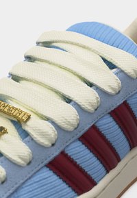 Κοντινό πλάνο μπλε αθλητικού παπουτσιού με χοντρές κρεμ κορδόνια, κόκκινες ρίγες και χρυσές άκρες κορδονιών, που δείχνει τις υφασμάτινες υφές και τις λεπτομέρειες ραφής.