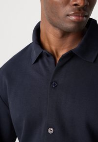 Camisa polo de color azul marino con cuello doblado, que presenta dos botones, fabricada con tejido suave, con una vista en primer plano del escote.