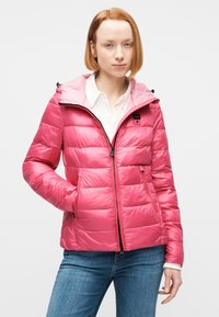 Νεαρή γυναίκα με κοντά κόκκινα μαλλιά, ντυμένη με ροζ μπουφάν τύπου puffer πάνω από λευκό πουκάμισο και μπλε τζιν, στέκεται με το ένα χέρι στην τσέπη.