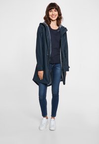 Giacca impermeabile navy con cappuccio, coulisse regolabile e foderato a righe. Abbinata a un top navy, jeans blue e sneakers bianche.