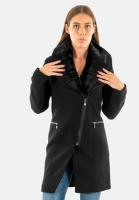 Manteau noir avec un col en fausse fourrure, fermeture éclair, deux poches avant zippées et silhouette allongée, porté sur un jean. Texture lisse.