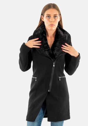 Manteau noir avec un col en fausse fourrure, fermeture éclair, deux poches avant zippées et silhouette allongée, porté sur un jean. Texture lisse.