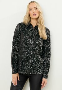 Schwarze Paillettenbluse mit kragenförmigem Ausschnitt, langen Ärmeln und einer strukturierten Oberfläche, die das Licht reflektiert, kombiniert mit schwarzen Lederhosen.