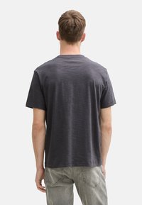 Camiseta gris de manga corta hecha de una tela suave, con cuello redondo y un corte relajado. Llevada con jeans de color claro. Se muestra vista trasera.