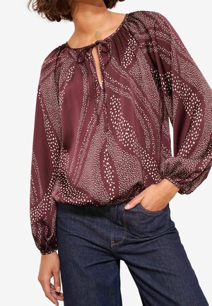 Vrouw draagt een lange mouwen bordeauxrode blouse met beige stippen en abstracte patronen, voorzien van een strik bij de halslijn, gecombineerd met donkere blauwe spijkerbroek.