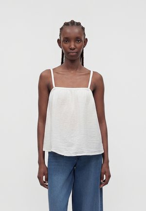 Γυναίκα με πλεξούδα, που φοράει άνετο λευκό αμάνικο τοπ και μπλε τζιν, στέκεται μπροστά σε απλό φωτεινό φόντο.