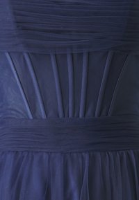 Rochie bleumarin cu pliuri stratificate și corset ajustat. Textură transparentă în partea de sus și material fluid și moale la fustă. Detalii cu cusături verticale.