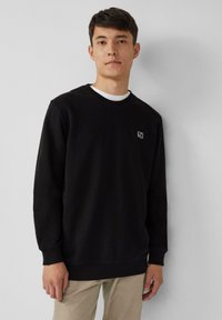 Giovane uomo in piedi contro un muro semplice, indossa un felpa nera con un piccolo logo bianco sul petto e pantaloni beige.