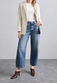 Kvinna som bär en krämfärgad kordflamsjacka, vit skjorta, vida blå jeans och bruna klackmules stående på ett grått golv.