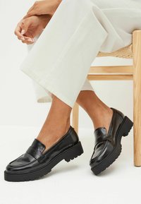 Mocassini in pelle nera con suola platform spessa, texture liscia e dettaglio a strappo sulla parte anteriore, abbinati a pantaloni bianchi.