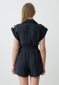 Zwarte short-sleeve jumpsuit met een kraag, strikceintuur en een zachte textuur. Heeft opgerolde mouwen en een aansluitend ontwerp met een relaxte pasvorm.