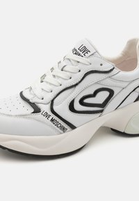 Hvite sneakers med et svart hjerte-design og merkevare. Glatt lær-overdel, snøring, og teksturert såle for grep.