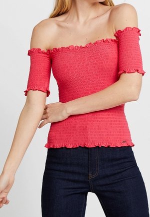 Mujer con un top rosa ajustado de hombros descubiertos con mangas cortas y bordes con volantes, combinado con vaqueros azul oscuro.