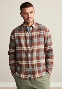 Camicia di flanella a quadri rossi, marroni e blu. Presenta un colletto con bottoni e cinque bottoni. Tessuto morbido con maniche lunghe, abbinata a una maglietta grigia.