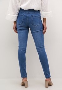 Mujer vestida con jeans ajustados azules, una blusa de manga larga blanca y zapatos de tacón cuadrado beige, de pie contra un fondo liso.