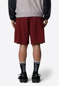 Cargo shorts in burgundy fabric, featuring side pockets, paired with black sneakers and striped socks displaying white text.