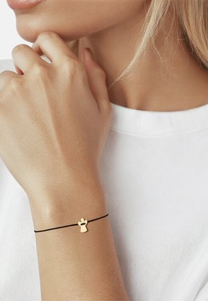 Frau trägt ein zartes schwarzes Kettenarmband mit einem kleinen goldenen Engelsanhänger am Handgelenk vor weißem Hemdhintergrund.