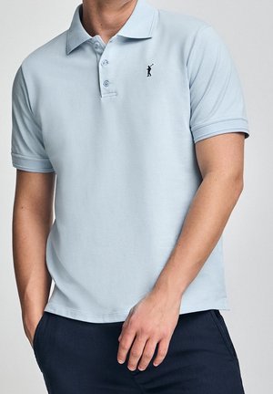 Męski tors w jasnoniebieskiej koszulce polo z krótkim rękawem, kołnierzykiem i małym czarnym haftowanym logo, w połączeniu z ciemnoniebieskimi spodniami.