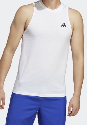 Mężczyzna ubrany w białą sportową koszulkę bez rękawów z czarnym logo i niebieskie szorty, stojący z rękami swobodnie opuszczonymi wzdłuż ciała.