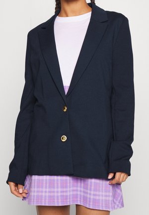 Femme portant un blazer bleu marine avec des boutons dorés sur un haut blanc et une jupe à carreaux violette, vue du cou jusqu'à mi-cuisse.