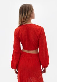 Top corto rosso con maniche lunghe a sbuffo, realizzato in tessuto testurizzato con motivi ad occhio. Gonna aderente coordinata con vita elastica.