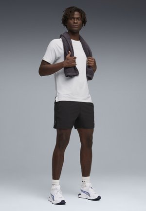 Mann trägt weißes T-Shirt, schwarze Shorts, weiße Turnschuhe, hält ein dunkles Handtuch um den Hals und steht vor grauem Hintergrund.