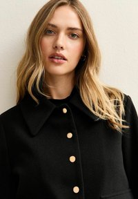 Czarny wełniany płaszcz z kołnierzem, z pięcioma dużymi beżowymi guzikami z przodu. Gładka faktura i dopasowany krój.