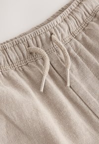 Primer plano de la cintura de unos pantalones de lino beige con elástico fruncido y cordones atados.