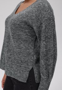 Grauer Strickpullover mit V-Ausschnitt, seitlichen Schlitzen und strukturierten Ärmeln. Weiches Material mit einem leicht lockeren Schnitt, kombiniert mit dunklen Jeans.