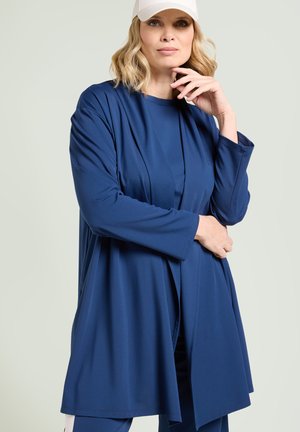Mujer rubia con gorra blanca y conjunto azul de manga larga posa con una mano junto a la cara y la otra sujetando su costado.
