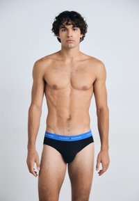 Schwarze Baumwollslips mit einem blauen elastischen Bund, der das "TOMMY HILFIGER"-Logo trägt. Das Modell hat eine schlanke Figur und steht aufrecht vor einem neutralen Hintergrund.